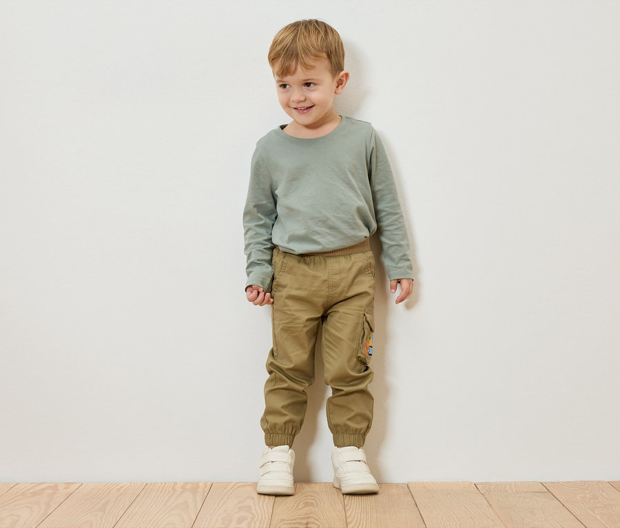 Chlapec s blond vlasmi stojí pred bielou stenou a má na sebe zelené tričko s dlhým rukávom, khaki nohavice a biele topánky.