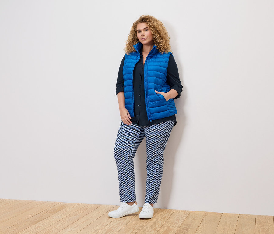 Žena s kučeravými vlasmi v modrej veste, tmavej košeli a vzorovaných nohaviciach stojí pri bielej stene.