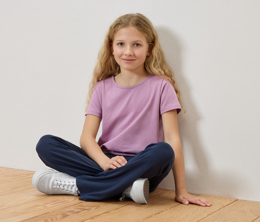 Dievča s blond vlasmi sedí so skríženými nohami na drevenej podlahe, má na sebe fialové tričko a modré nohavice.