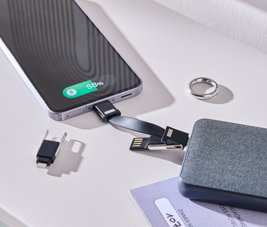 Smartfón sa nabíja pomocou kábla a sivej batérie na bielom stole. Vedľa leží prsteň a izbová karta.