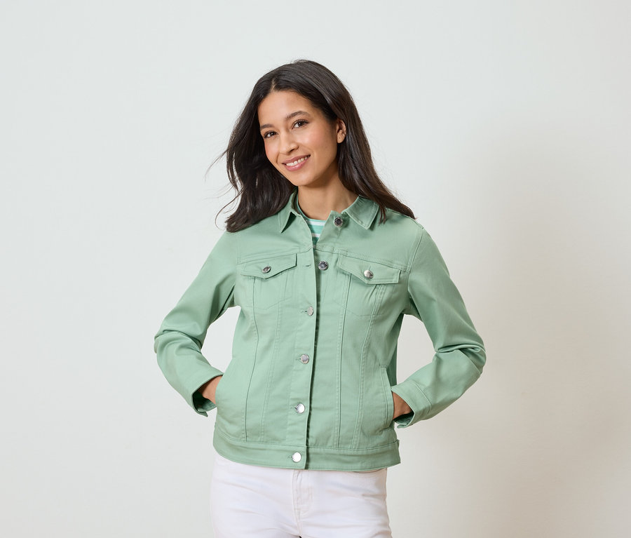 Žena pózuje s rukami vo vreckách, oblečená v svetlozelenej džínsovej bunde a bielych nohaviciach.