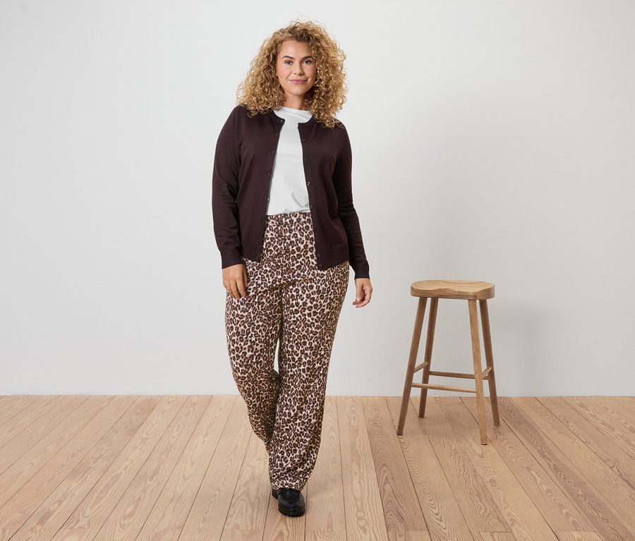 Žena s kučeravými vlasmi má na sebe hnedý kardigán, biele tričko a nohavice s leopardím vzorom.