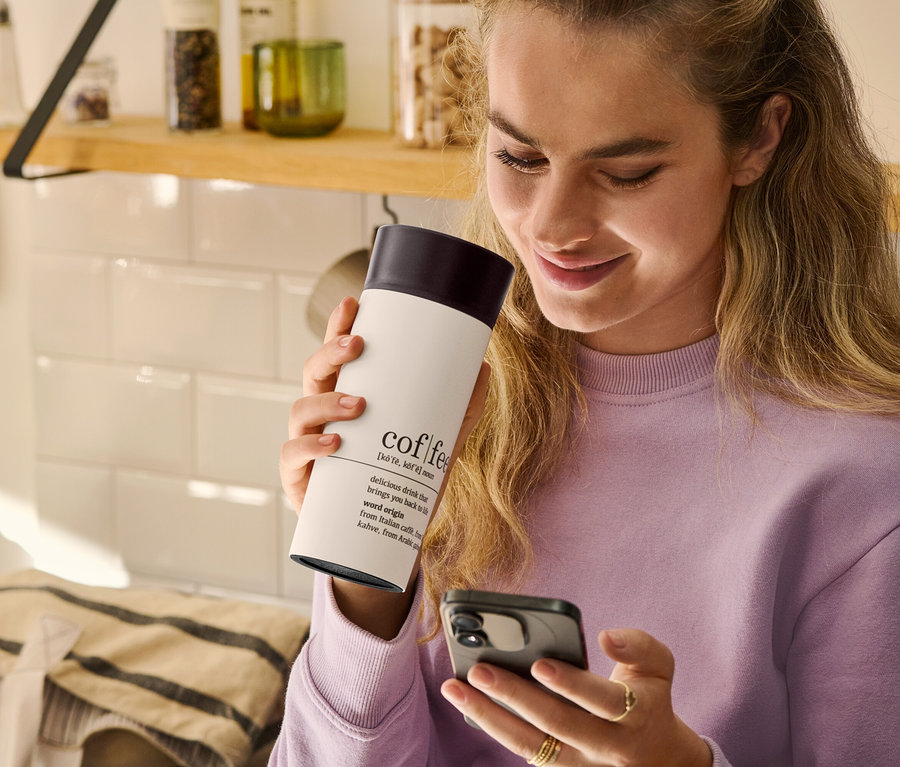 Žena drží v ruke hrnček na kávu To Go, Coffee Dictionary a pozerá sa na mobilný telefón.