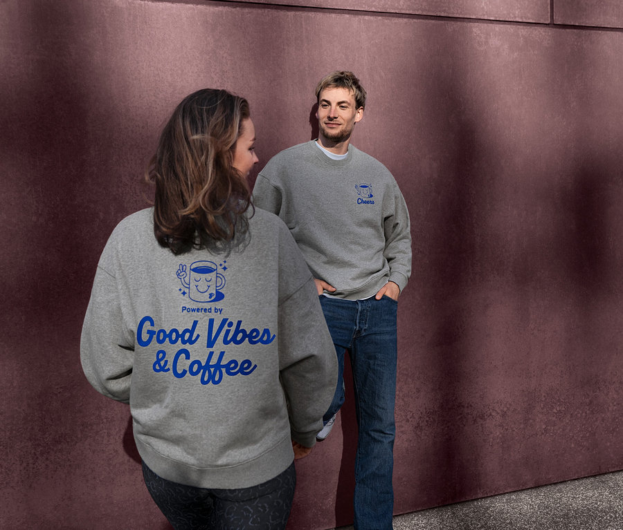 Žena a muž v sivých unisex mikinách Coffee Crew. Na chrbte mikiny ženy je modrý nápis "Powered by Good Vibes & Coffee". Muž sa pozerá na ženu, jeho mikina má modrý nápis "Cheers".