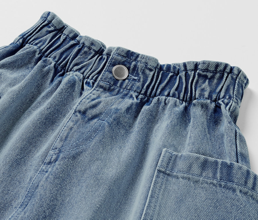 Detail detskej džínsovej sukne v štýle paperbag so zapínaním na gombík a vreckami.