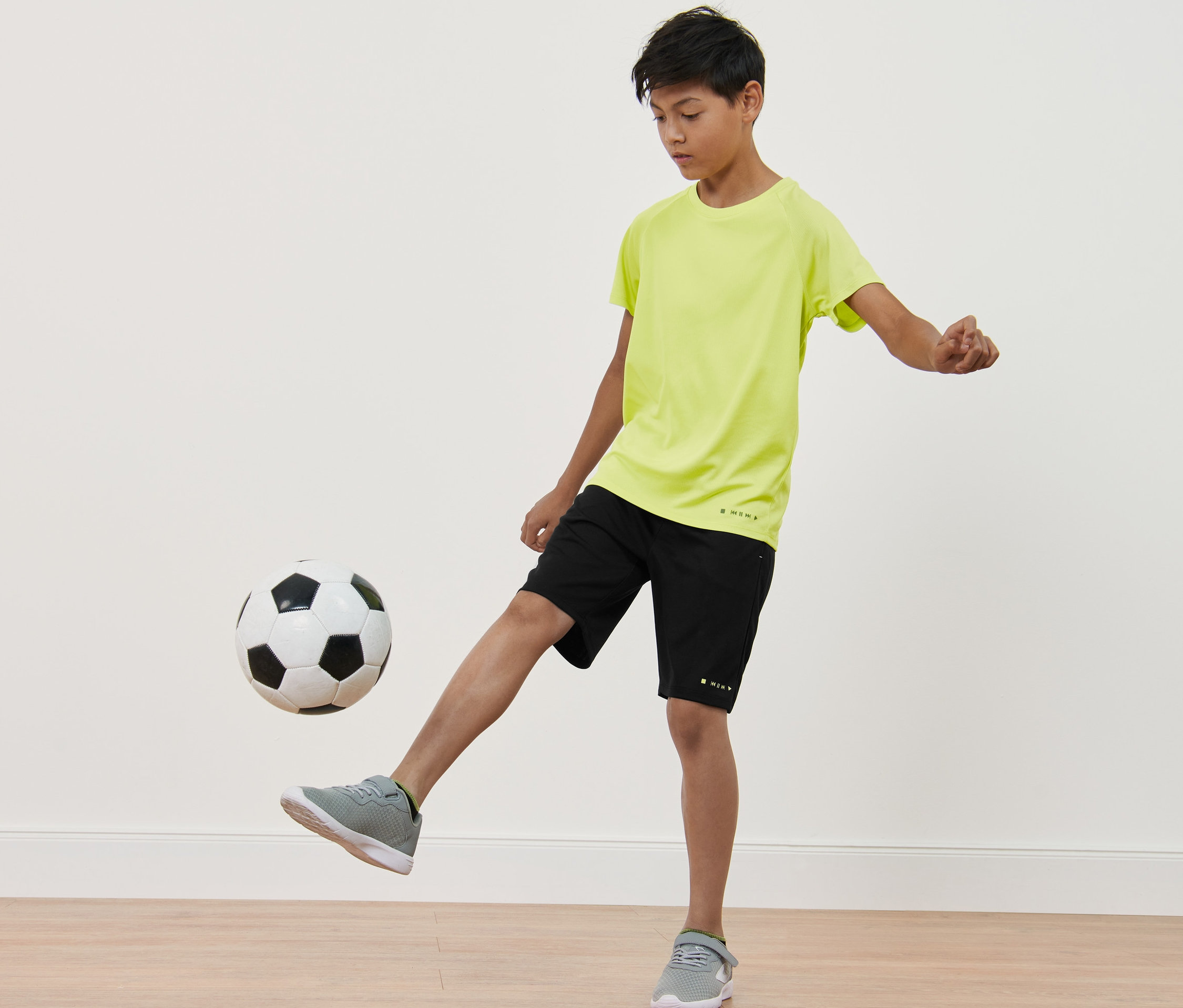 Chlapec kopne do futbalovej lopty. Má na sebe čierne športové šortky a neónovožlté funkčné tričko. Detské funkčné tričká, 2 ks.