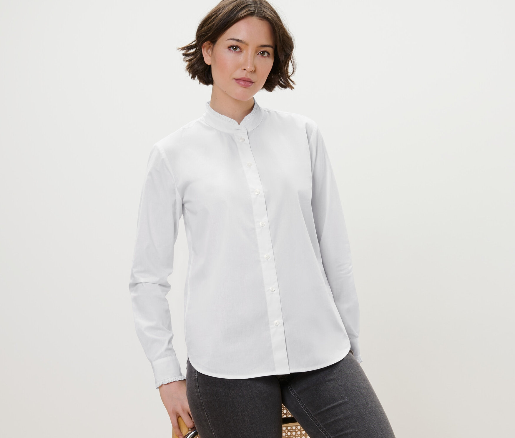 Žena pózuje na stoličke a má na sebe bielu blúzku s nariaseným detailom a sivé džínsy.