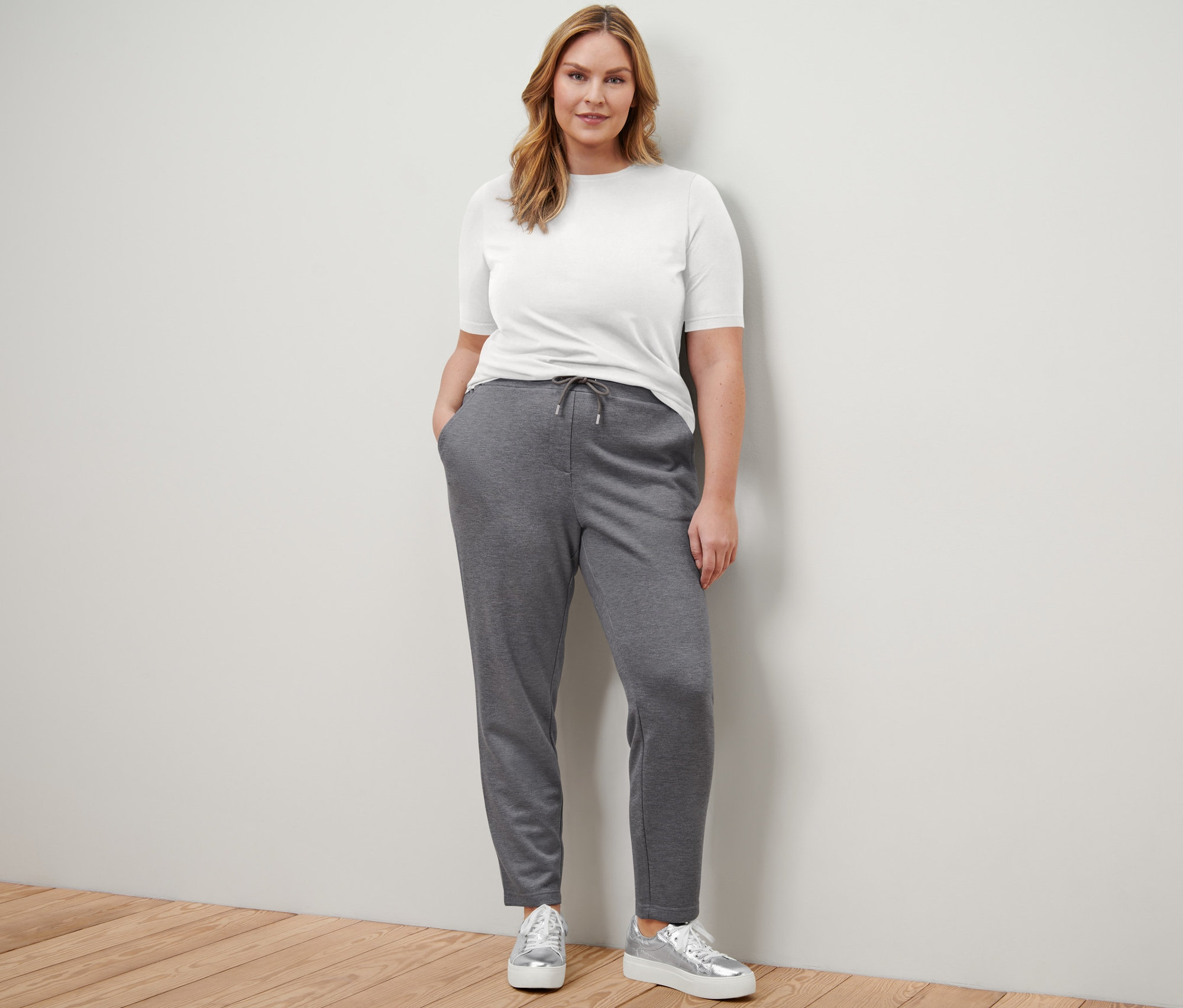 Žena stojí a má na sebe biele jednoduché tričko s polodlhým rukávom, tmavomodré jednoduché tričko s polodlhým rukávom, tepláky a tenisky na platforme z kože.