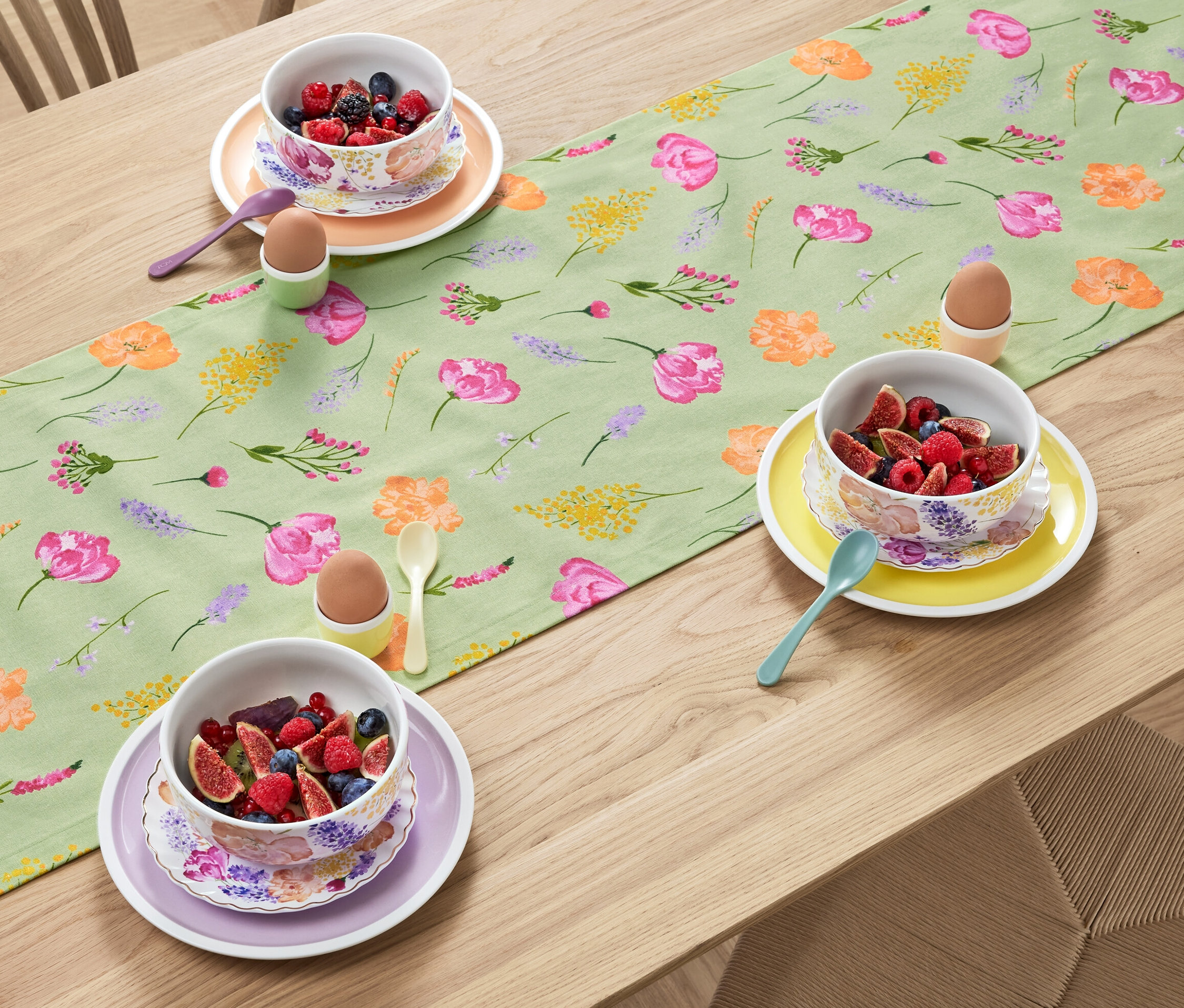 Na drevenom stole leží šalviový behúň s kvetinovou potlačou, na ktorom sú misky s ovocím, vajíčka v držiakoch a lyžičky.