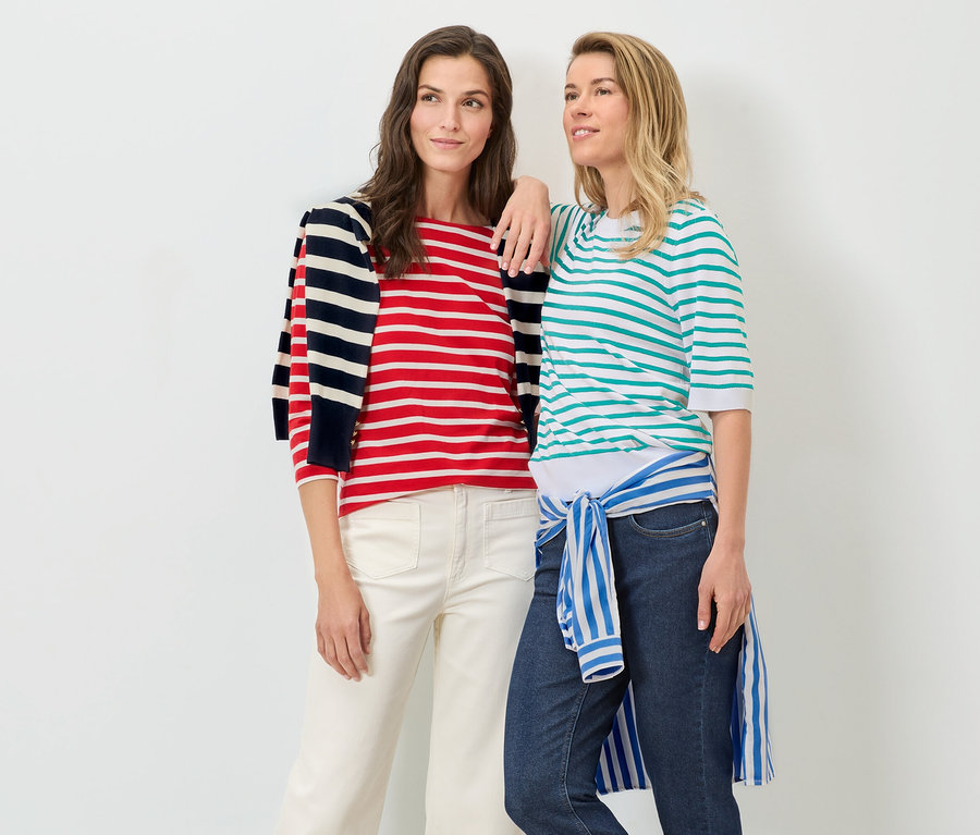 Dve ženy pózujú na bielom pozadí. Jedna má na sebe akvamarínový pruhovaný pulóver z jemného úpletu a džínsy, druhá má na sebe červeno-biely pruhovaný pulóver a biele nohavice.