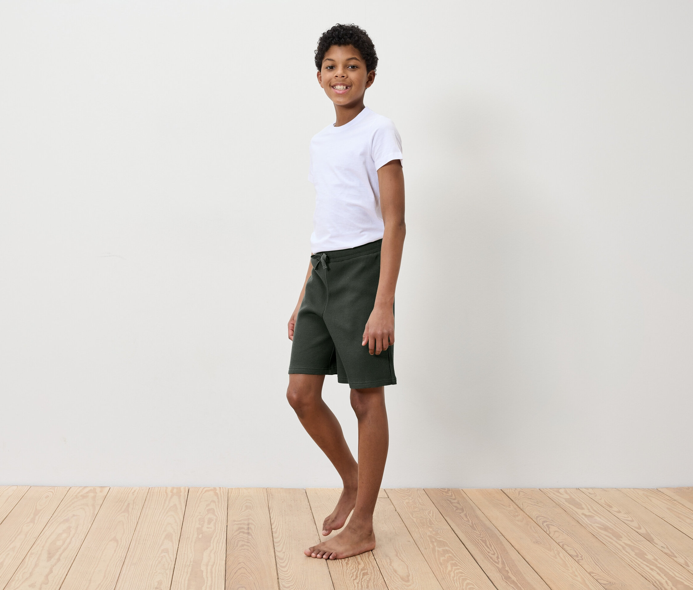 Chlapec v bielom tričku a zelených šortkách stojí na drevenej podlahe pred bielou stenou.