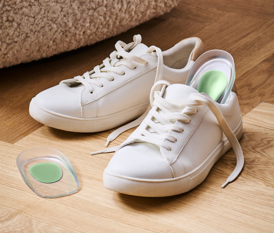 Dve biele tenisky ležia na drevenej podlahe, jedna so zelenou vložkou.