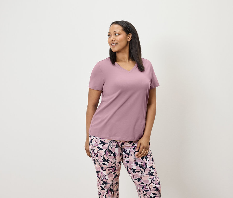 Žena v lila tričku s krátkym rukávom a kvetinových nohaviciach sa pozerá doprava.