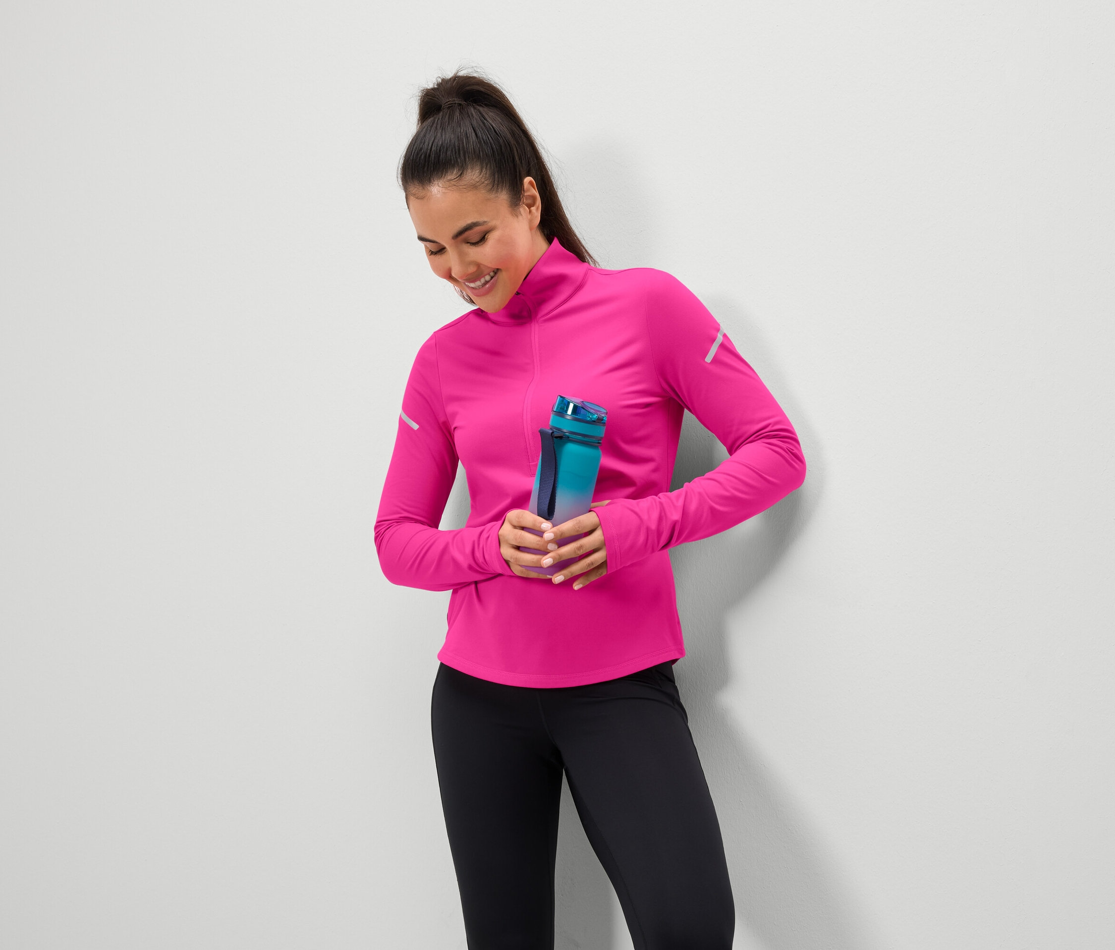 Usmievajúca sa žena drží modrozelenú fľašu na vodu, má na sebe ružový top a čierne legíny.