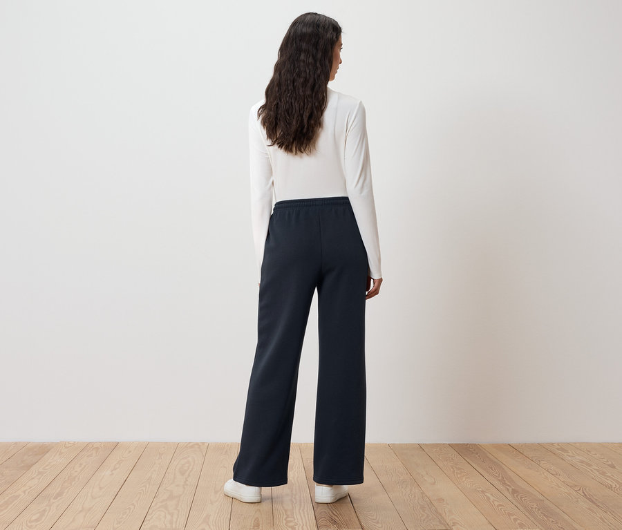 Žena s dlhými vlnitými vlasmi stojí chrbtom a má na sebe biely top s dlhým rukávom a tmavomodré široké nohavice.