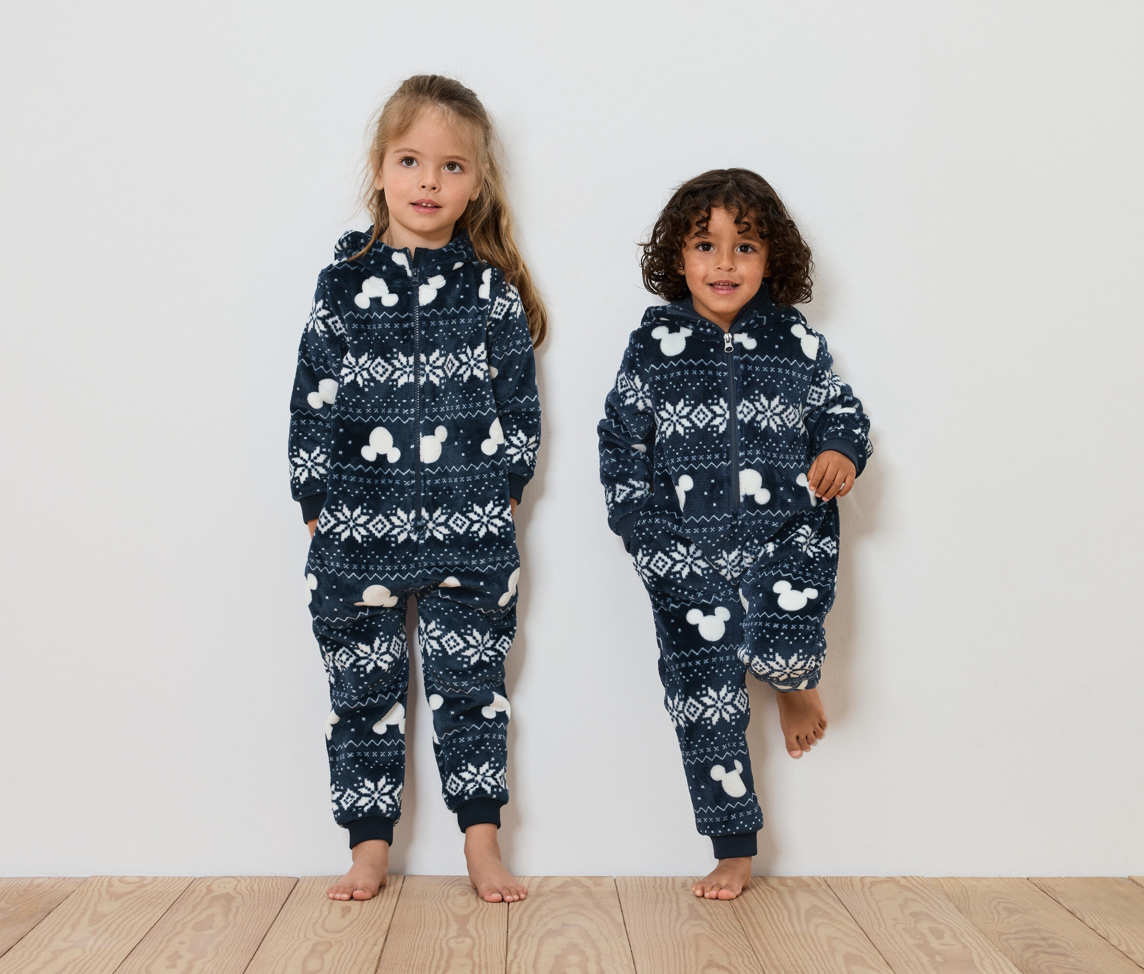 Dve deti stoja vedľa seba a majú na sebe modré vianočné overaly s kapucňou s motívmi Mickey Mouse.