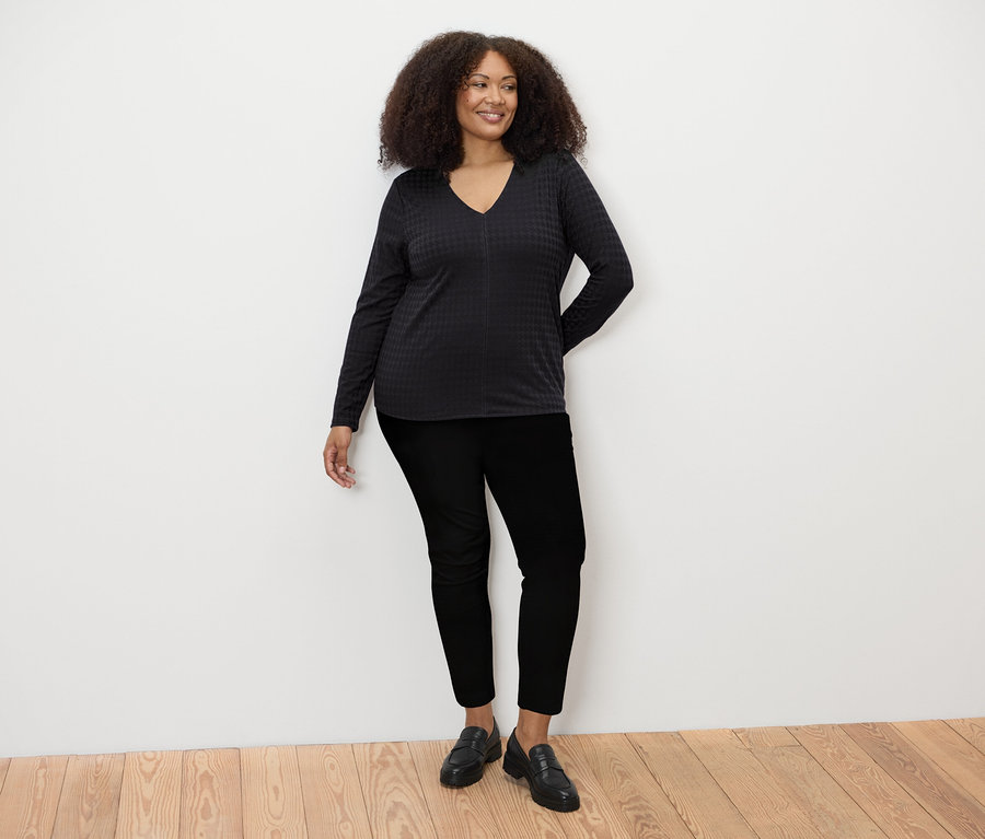Žena stojí a má na sebe čierne štruktúrované tričko s dlhým rukávom, čierne nohavice a čierne topánky.