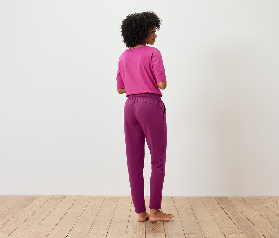 Žena s afro účesom stojí chrbtom a má na sebe ružové tričko a fialové nohavice.