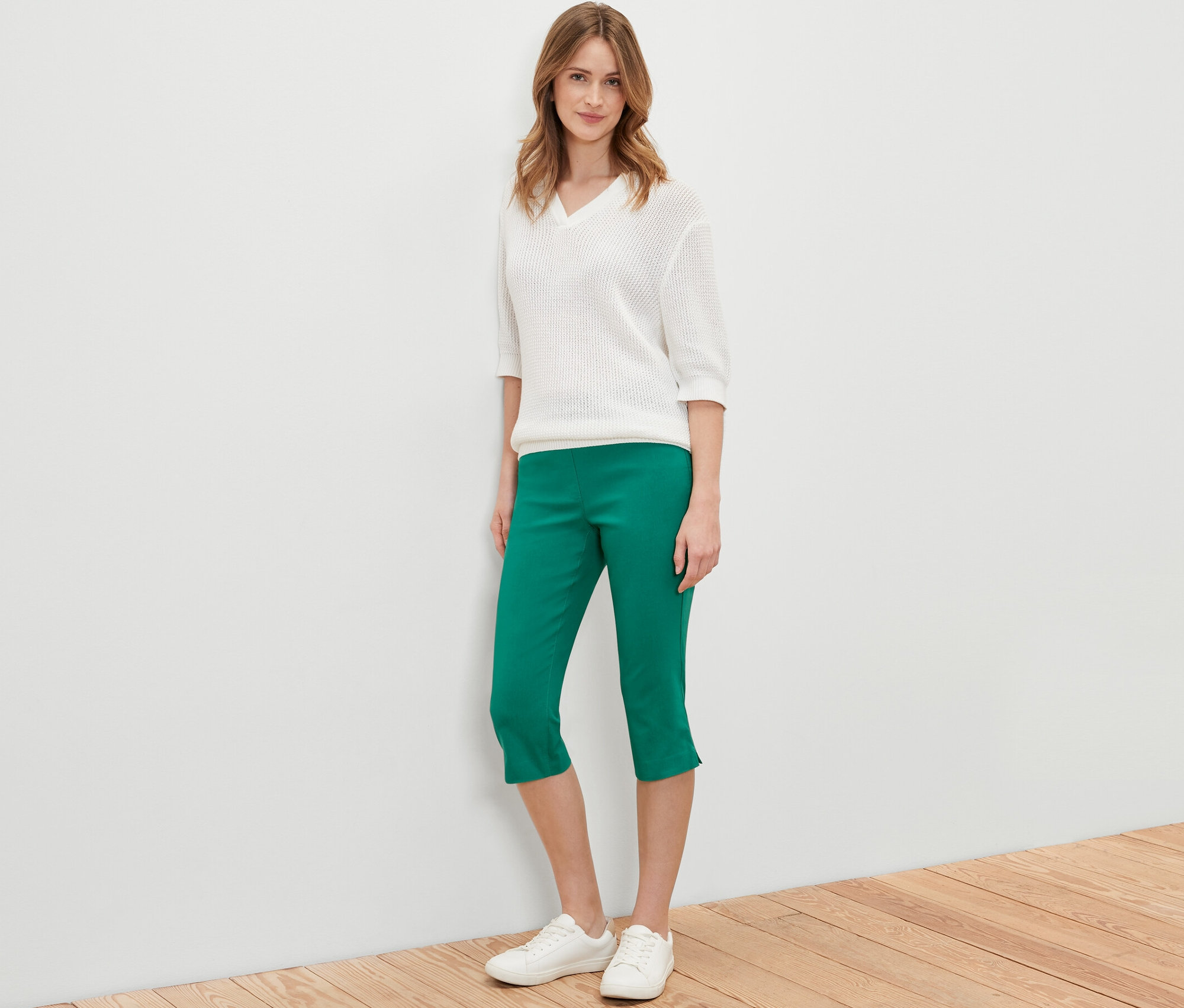 Žena stojí a pózuje v zelených 3/4 bengalínových nohaviciach, bielom svetri a bielych topánkach.