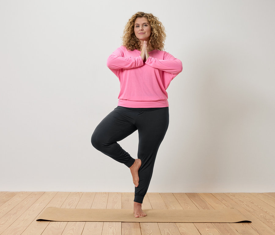 Žena s kučeravými vlasmi cvičí jogu na béžovej podložke, oblečená v ružovom tope a sivých nohaviciach.