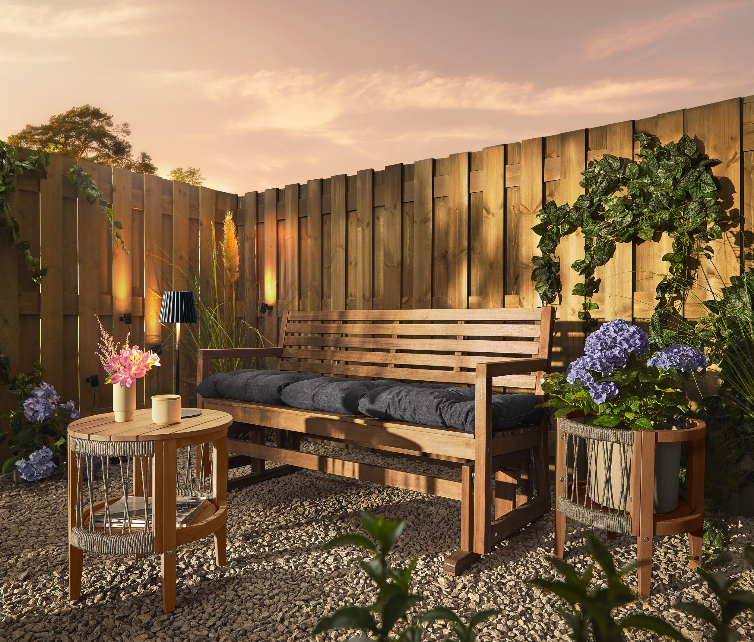 Drevená lavica s vankúšmi stojí na štrkovom podklade pred dreveným plotom. Na plote sú upevnené 3 solárne svietidlá na strešné žľaby.
