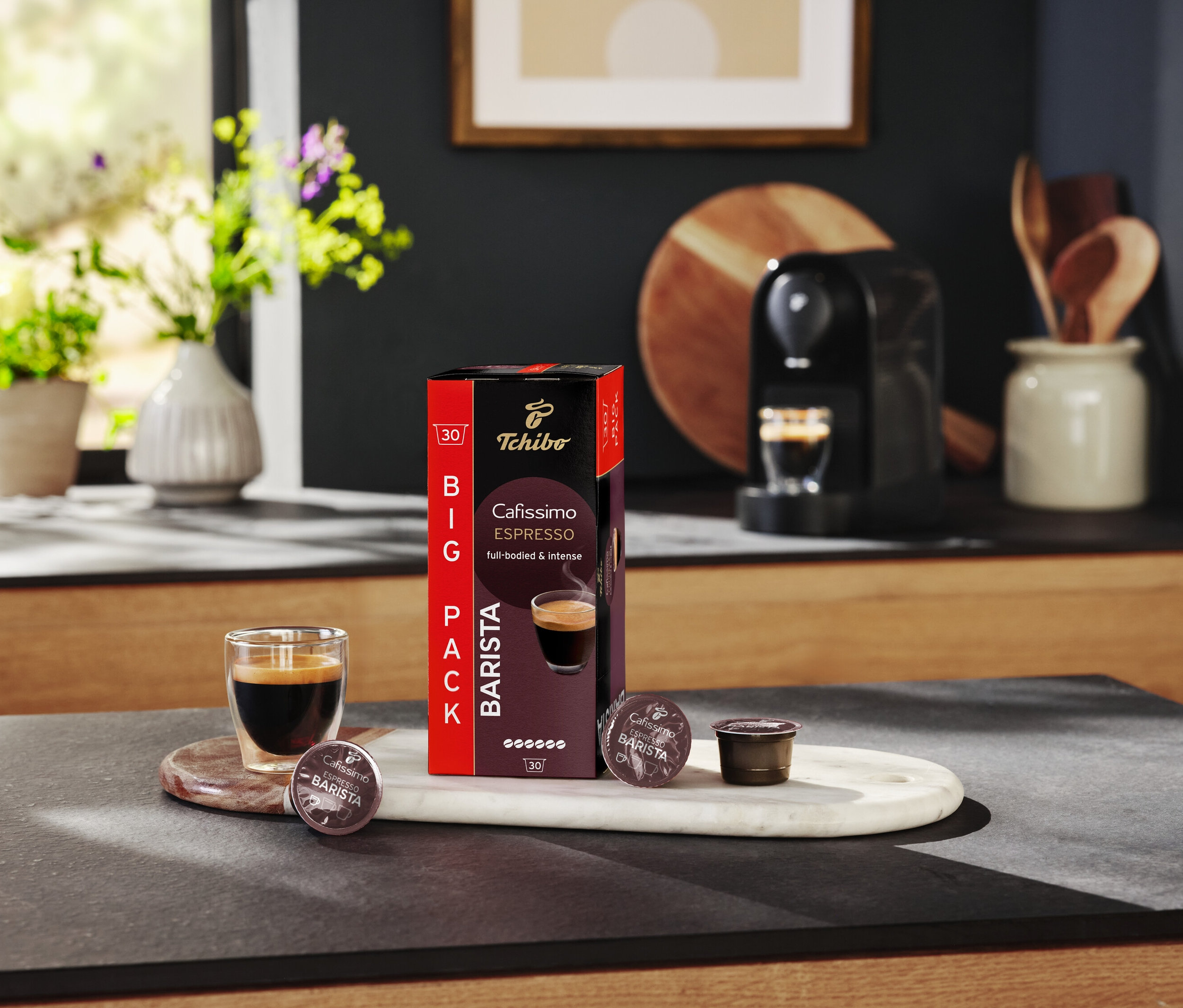 Šálka espressa stojí vedľa balenia kapsúl Tchibo Cafissimo Barista na kuchynskom stole.