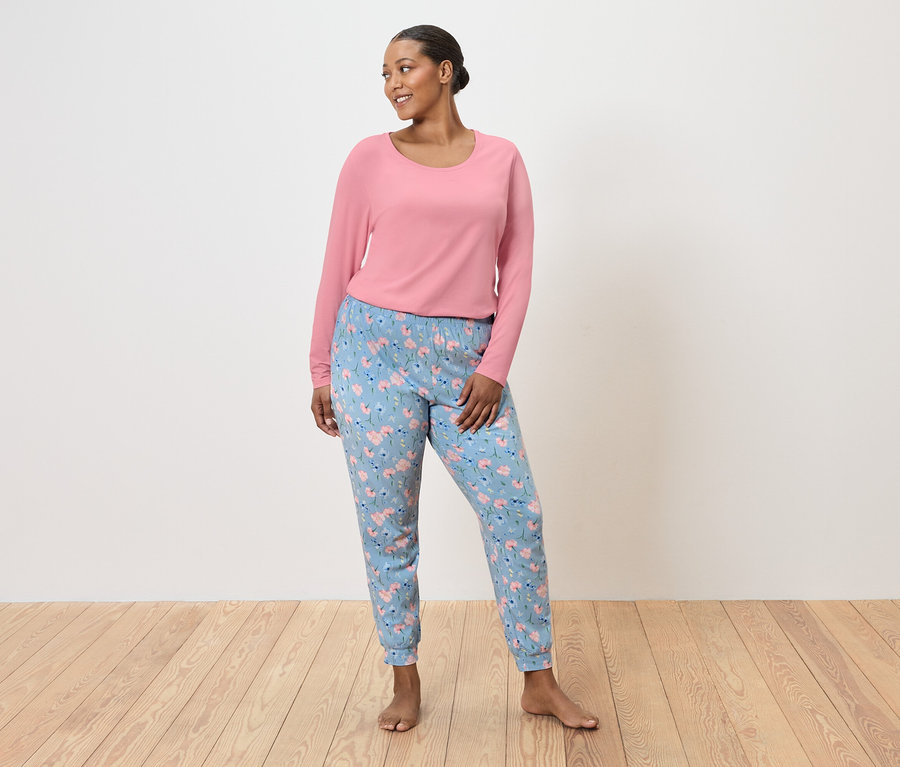 Žena stojí naboso, má na sebe ružový sveter a svetlomodré nohavice s kvetinovým vzorom.