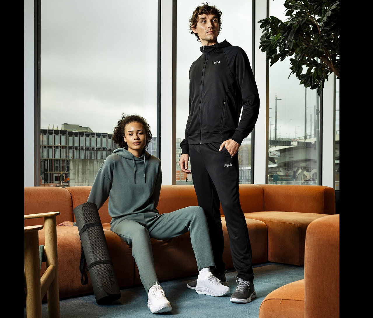 Mladá žena sedí na oranžovej pohovke a drží čiernu podložku na jogu. Vedľa nej stojí mladý muž v čiernej teplákovej súprave.