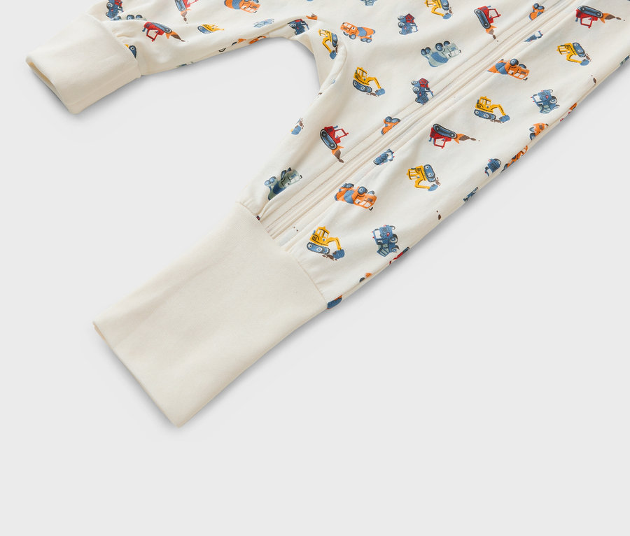 Detailný záber na dojčenské pyžamo na zips so vzorom rôznych stavebných vozidiel na béžovom pozadí.
