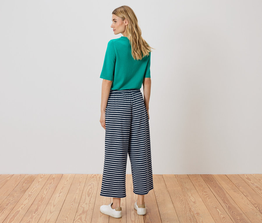 Žena stojí chrbtom k fotoaparátu, má na sebe zelený top a námornícko-biele pásikavé culottes nohavice.