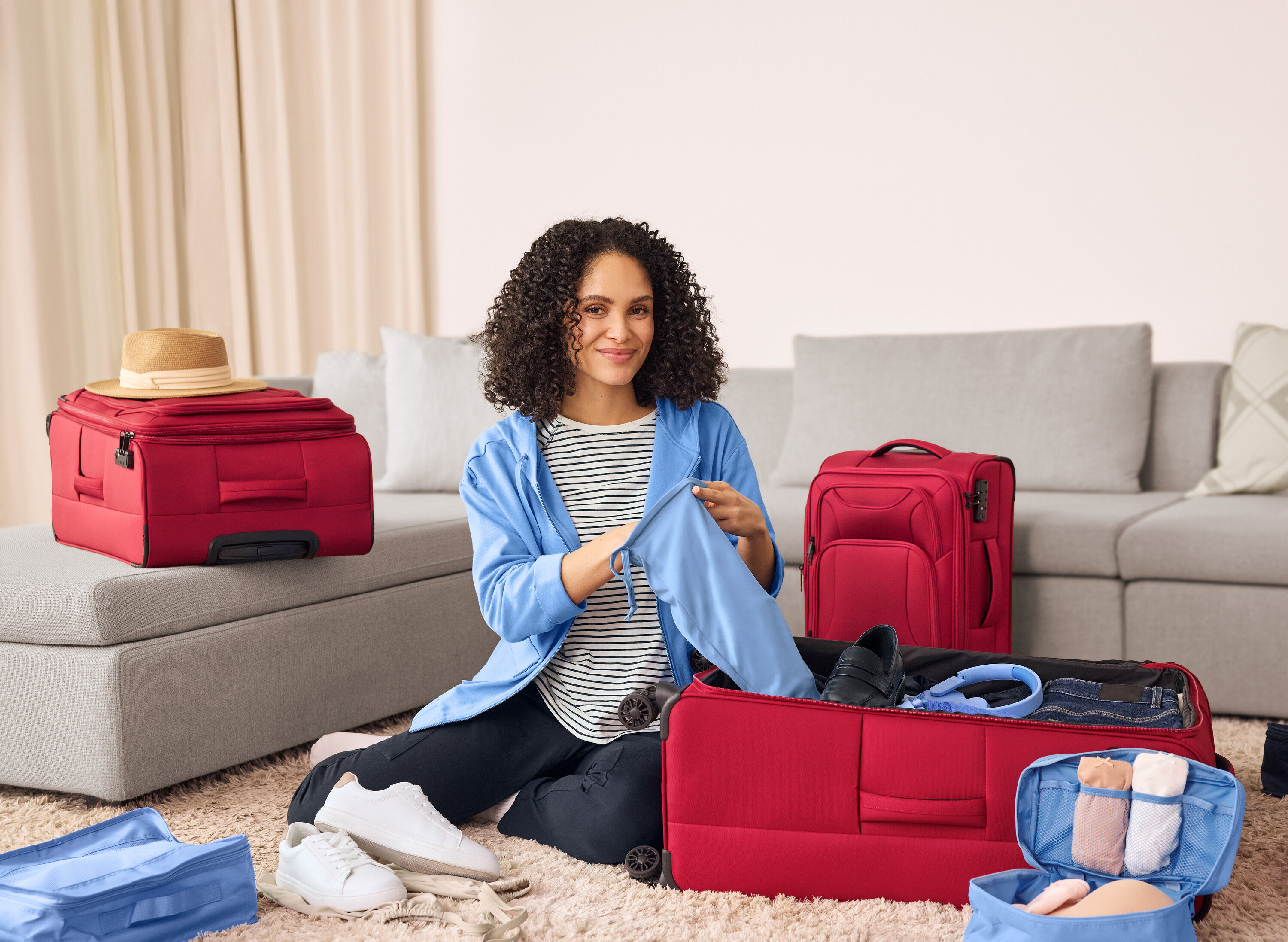 Žena sedí na zemi a balí tmavomodré tepláky s vreckami cargo do červeného kufra. Vedľa nej sú kožené tenisky.