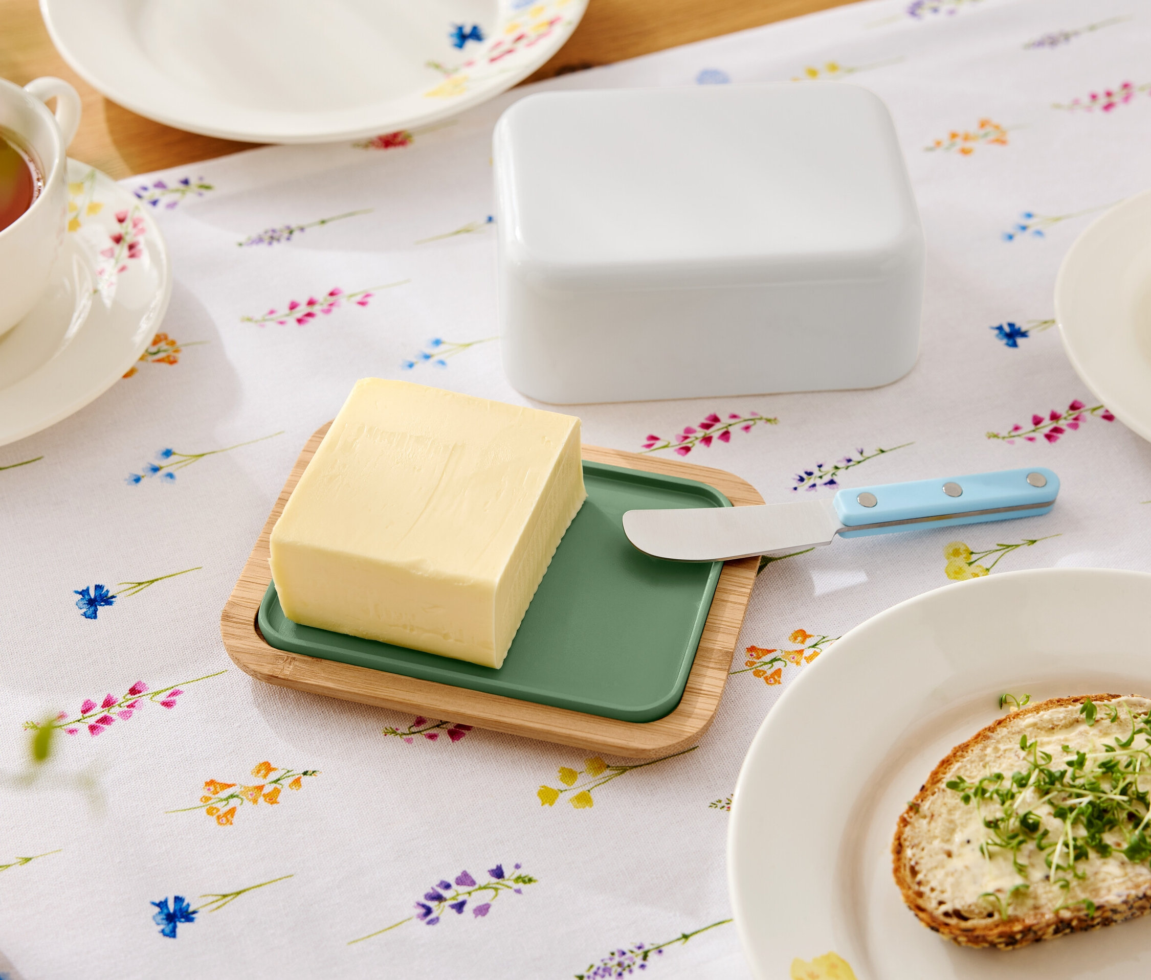 Na kvetinovom stole leží maslo na drevenom tanieri so zelenou vložkou, nôž na maslo, maselnička, šálka čaju a tanier so sendvičom s maslom.