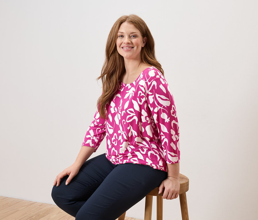 Žena s dlhými, ryšavými vlasmi sedí na drevenej stoličke a má na sebe ružovú blúzku s bielymi listami a tmavomodré nohavice.