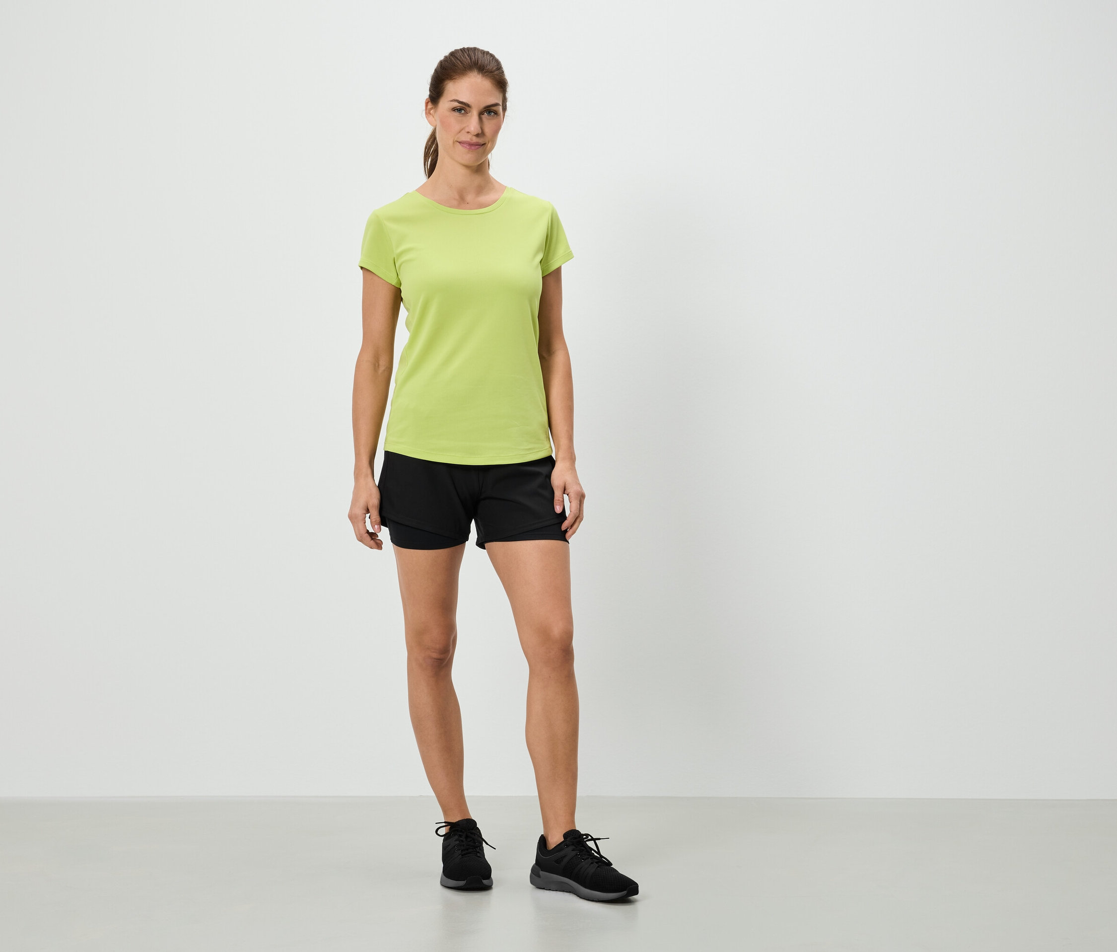 Žena stojí v svetlozelenom tričku, čiernych šortkách a čiernych teniskách pred bielou stenou.