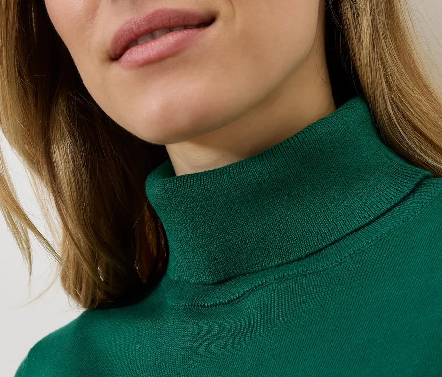 Detailný záber ženy s blond vlasmi v smaragdovom roláku.