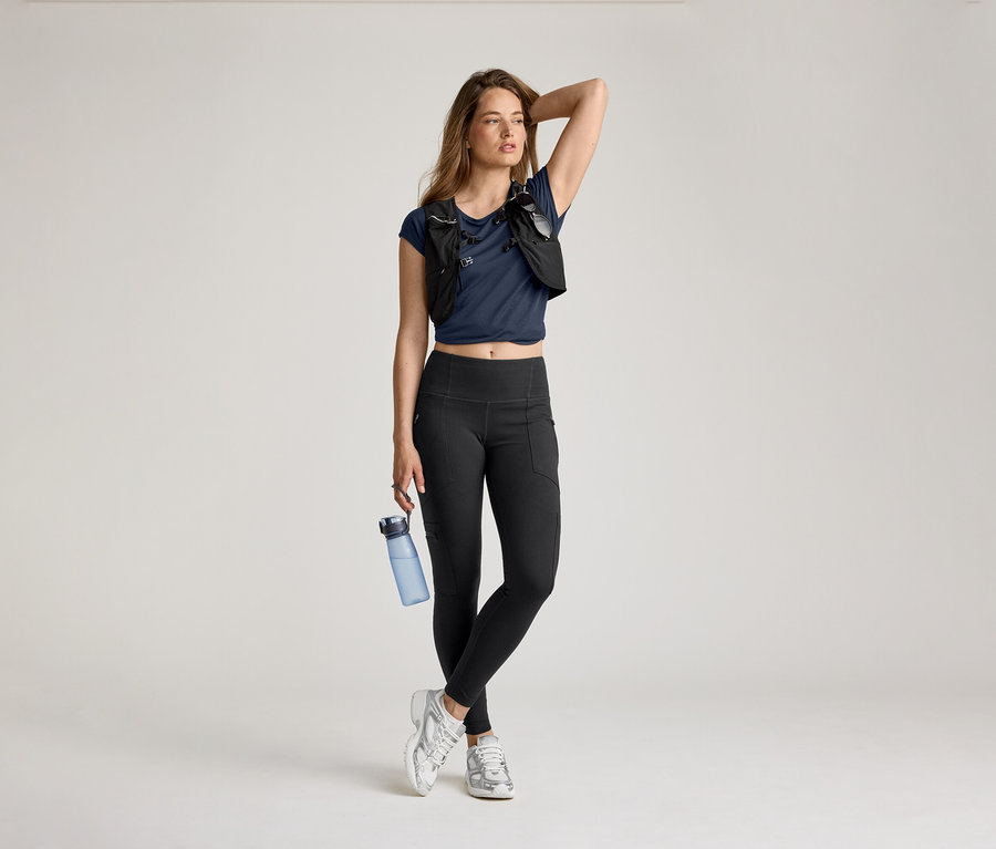 Žena v outdoorových legínsach, modrom tričku a bežeckej veste pózuje s fľašou vody a rukou na hlave.