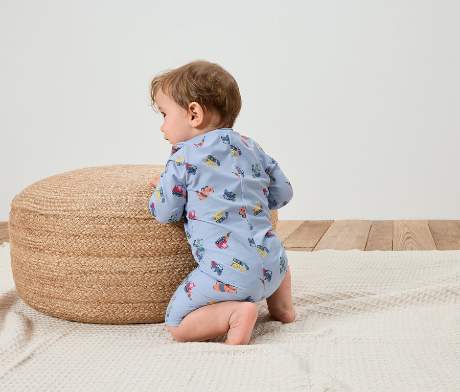 Dieťa v svetlomodrom overale so vzorom stavebných vozidiel kľačí na bielej deke vedľa okrúhleho pleteného pufu.
