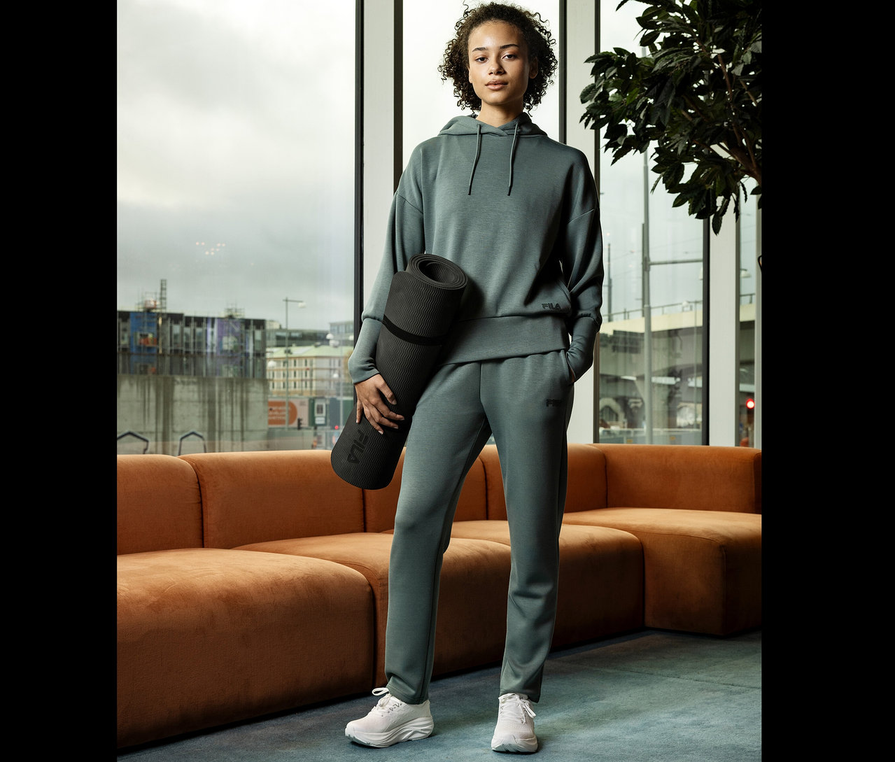 Žena v zelenej teplákovej súprave drží podložku na jogu pred oknom.