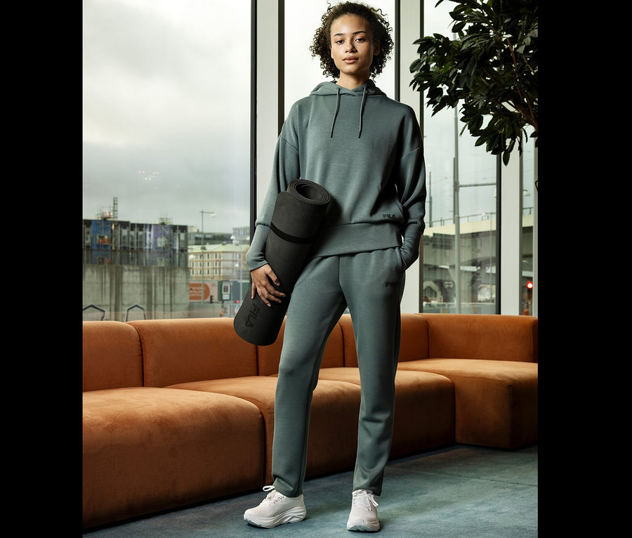 Žena v zelenej teplákovej súprave drží podložku na jogu pred oknom.