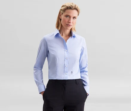 Žena má na sebe svetlomodrú blúzku Seidensticker Slim Fit non iron a čierne nohavice, ruky má vo vreckách.