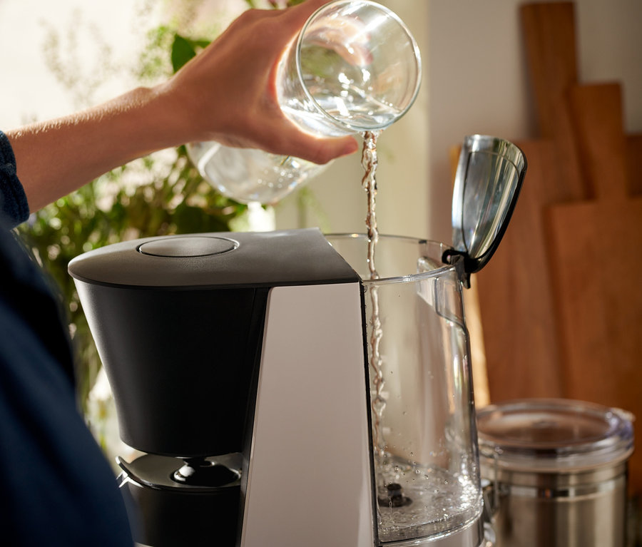 Žena naleva vodu z priehľadného pohára do sivo-čierneho kávovaru na prekvapkávanú kávu »Let's Brew«.