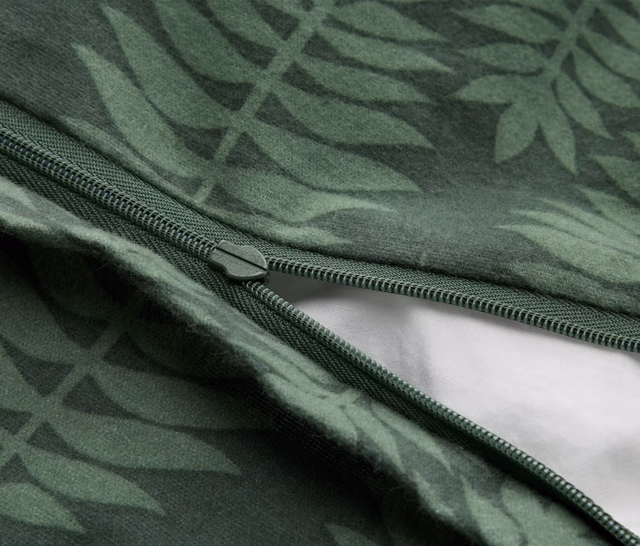 Detail tmavozelenej látky so svetlozeleným listovým vzorom. Zelený zips je čiastočne otvorený a odhaľuje biely materiál pod ním.