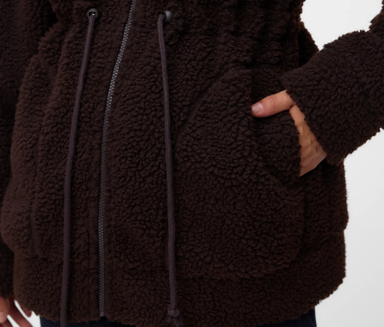 Detail osoby v hnedej, huňatej bunde s rukou vo vrecku. Bunda má zips a šnúrky na stiahnutie v páse.