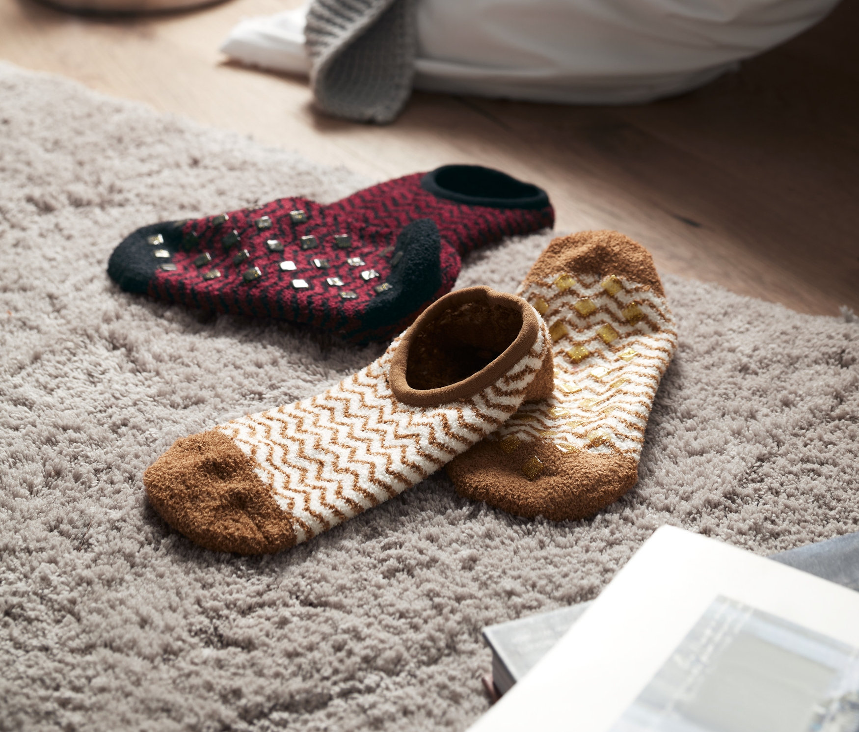 Dva páry ponožkových papúč ležia na sivom koberci, v pozadí sviečky a deka.