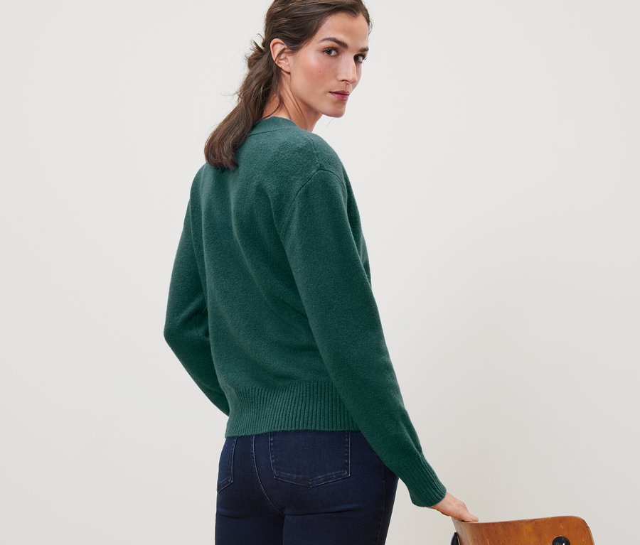 Žena sa pozerá cez plece a má na sebe tmavozelený pletený sveter s vlnou a džínsy.