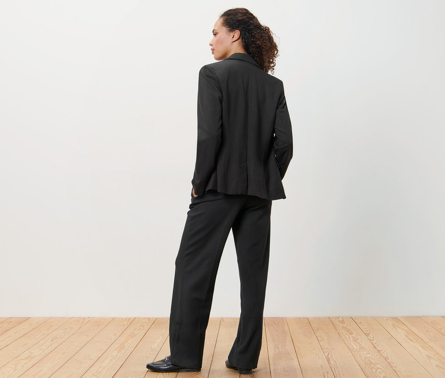 Žena v čiernom džersejovom saku a nohaviciach stojí chrbtom ku kamere.