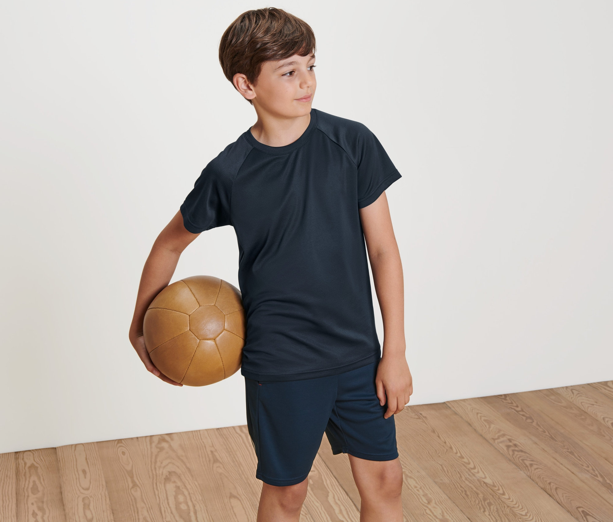 Chlapec drží hnedú koženú loptu. Má na sebe jedno z dvoch detských funkčných tričiek.