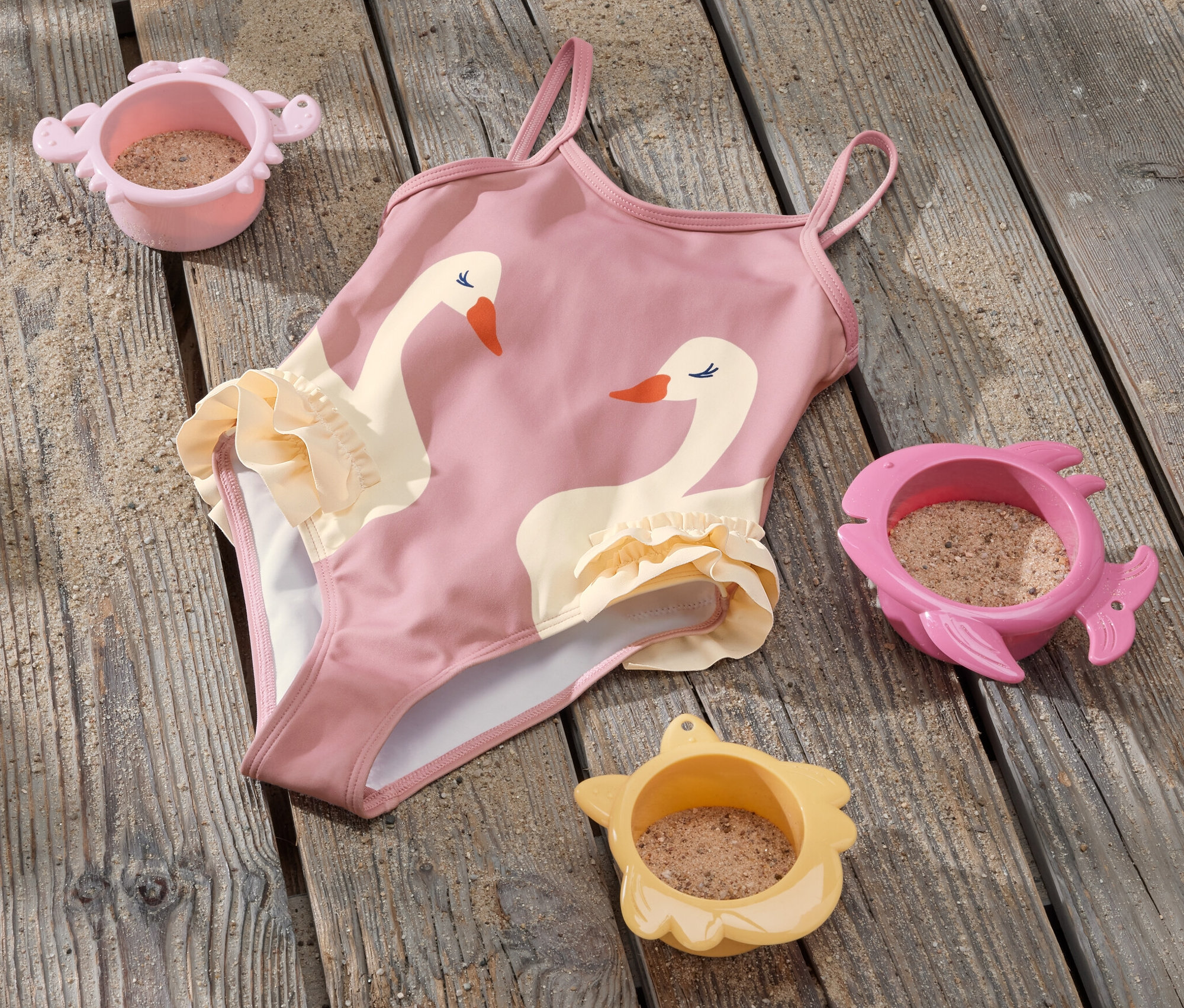 Detské jednodielne plavky s volánikmi a motívom labute ležia na drevenej podlahe vedľa troch hračiek do piesku.