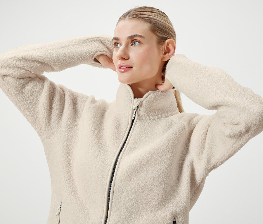 Žena s blond vlasmi stiahnutými do chvosta pózuje v krémovej fleecovej bunde na zips.