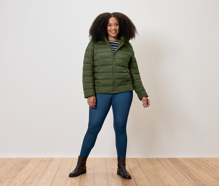Žena stojí a má na sebe olivovozelenú prešívanú bundu, džínsy a hnedé topánky.